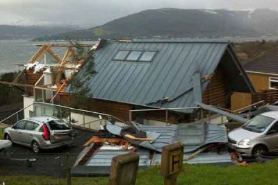 A house with part of its roof torn off and broken timber showing, with two cars parked in front